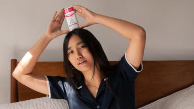 Woman with long black hair wearing navy pajamas and sitting up in bed holding a Schmidt's Rose+Vanilla natural deodorant over her head